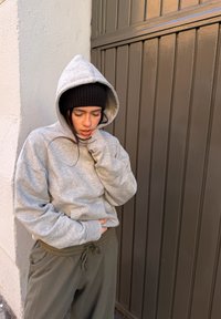 Jeune personne portant un sweat à capuche gris et un bonnet noir, appuyée contre un mur, tenant son ventre avec une expression de douleur.