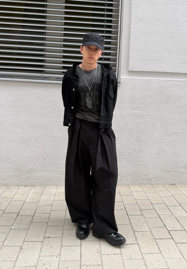 Jeune personne portant une casquette noire, des colliers superposés, une veste noire, un pantalon noir surdimensionné et des chaussures noires, debout contre un mur blanc.