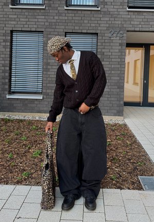 Schwarze Strickjacke über einem weißen Hemd und einer gemusterten Krawatte, weite schwarze Hosen und eine Handtasche mit Leopardenmuster.