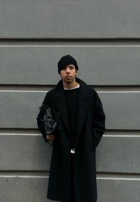 Abrigo negro texturizado llevado sobre un suéter negro, combinado con una camisa blanca. Sosteniendo un bolso negro. El fondo es una pared gris.