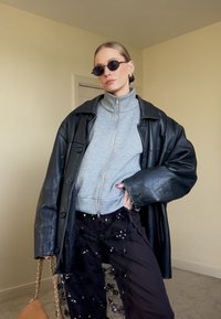 Femme portant une veste en cuir noire sur un haut à fermeture éclair gris, un pantalon noir transparent orné de paillettes, de petites lunettes de soleil noires, et tenant un sac à main beige avec une chaîne dorée.