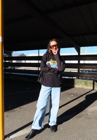 Čierna dlhá rukávová košeľa s grafickým dizajnom, svetlomodré široké nohavice, čierne mokasíny a nadrozmerné žlté slnečné okuliare. Stojí na parkovisku.