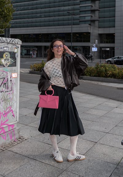 Falda negra plisada, blusa blanca de encaje, chaqueta bombers marrón de gran tamaño, bolso rosa, zapatillas blancas con rayas negras, gafas transparentes, fondo urbano.