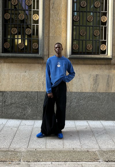 Sudadera azul de manga larga con logotipo gráfico y pantalones anchos negros, combinados con calzado azul, de pie frente a una pared de piedra con ventanas con patrones.