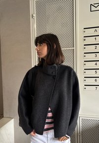 Tânără cu păr castaniu până la umeri poartă o jachetă gri închis cu nasturi, cămașă în dungi și pantaloni albi, stând lângă cutii poștale numerotate.