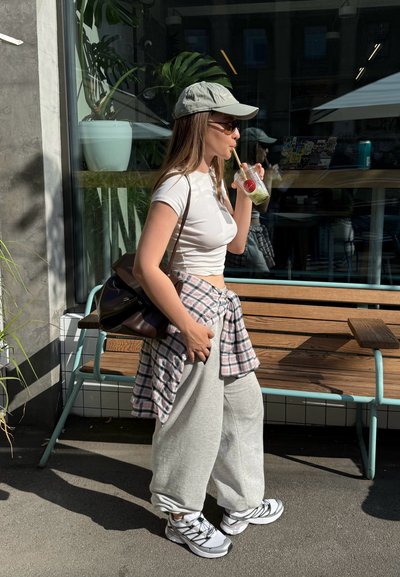 Outfit casual que incluye un top corto ajustado blanco, pantalones de chándal anchos gris claro, una camisa de franela a cuadros atada a la cintura y una gorra gris.