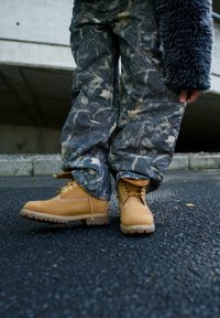 Bottes en cuir tan avec des semelles rugged, dotées de lacets jaunes et d'accents en maille, associées à un pantalon en camouflage gris à motifs, ample.