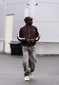 Homme marchant à l'extérieur portant une veste de survêtement marron, une casquette marron avec des cache-oreilles, un jean gris ample avec des porte-clés, et des baskets blanches.