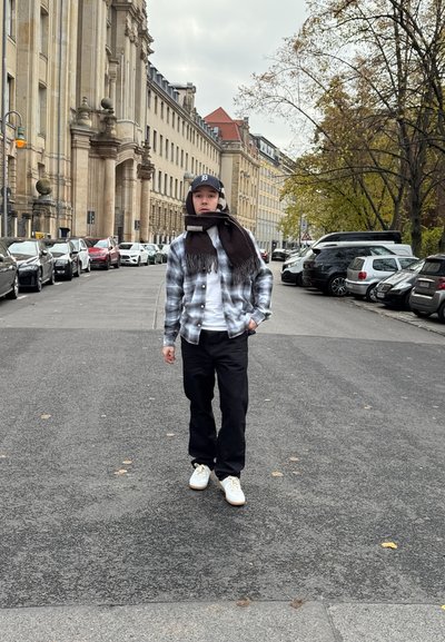 Camisa de franela de cuadros en azul y gris, camiseta blanca, pantalones negros, zapatillas blancas y una bufanda oscura. Entorno urbano con coches aparcados.