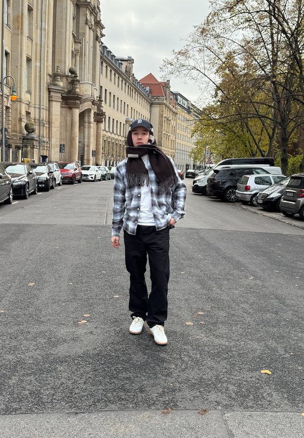 Camisa de franela de cuadros en azul y gris, camiseta blanca, pantalones negros, zapatillas blancas y una bufanda oscura. Entorno urbano con coches aparcados.
