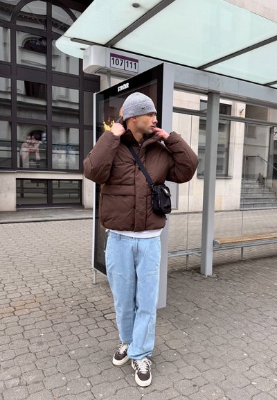 Chaqueta de plumas marrón, jeans celestes, gorro de punto gris. Bolsa de bandolera negra. De pie en una parada de autobús en una calle empedrada.
