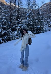 Femme portant une écharpe blanche et des moufles, debout dans une neige profonde près de pins couverts de neige, dans un paysage montagneux hivernal.