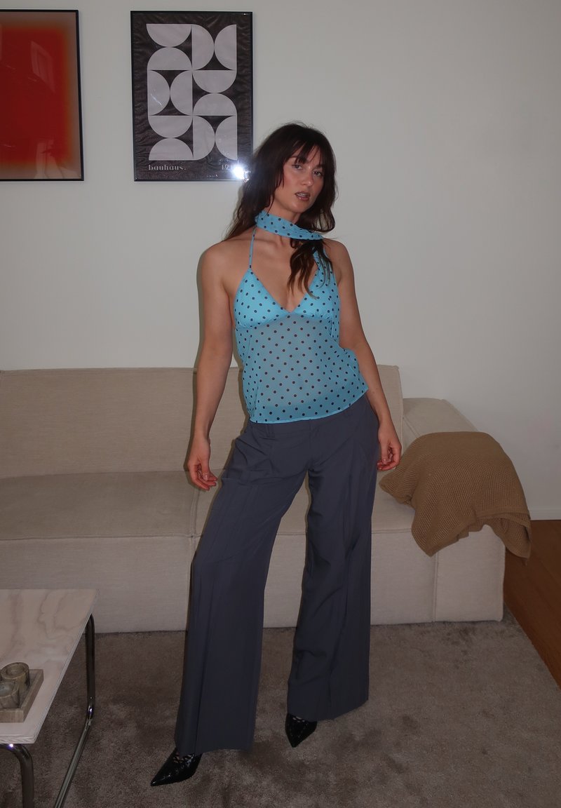 Jeune femme en haut bleu à pois, pantalon gris à jambes larges et talons noirs, debout dans un salon avec canapé et œuvre murale abstraite.