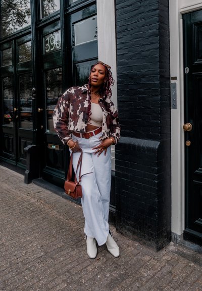 Chaqueta de patrón marrón y blanco sobre una camiseta beige, pantalones anchos blancos y botas de tobillo blancas. Bolsa de hombro marrón, los accesorios incluyen collares.