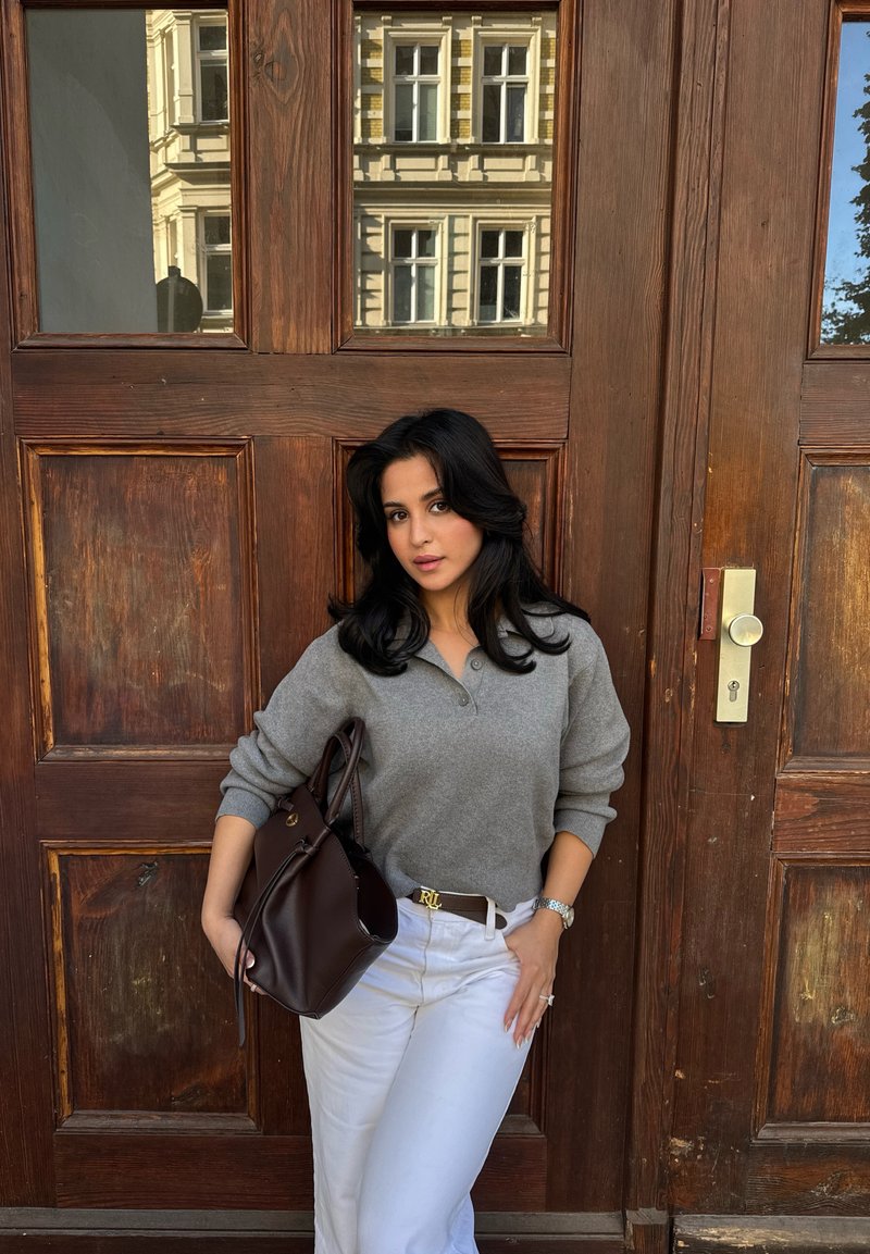Une femme en pull gris et pantalon blanc pose avec un sac à main marron devant une grande porte en bois.