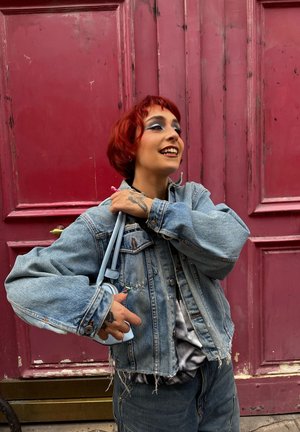 Joven con cabello corto y rojo y maquillaje, vistiendo una chaqueta de mezclilla desgastada, sosteniendo un pequeño bolso azul claro, de pie junto a una puerta roja.