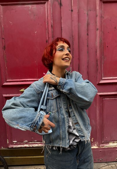 Joven con cabello corto y rojo y maquillaje, vistiendo una chaqueta de mezclilla desgastada, sosteniendo un pequeño bolso azul claro, de pie junto a una puerta roja.