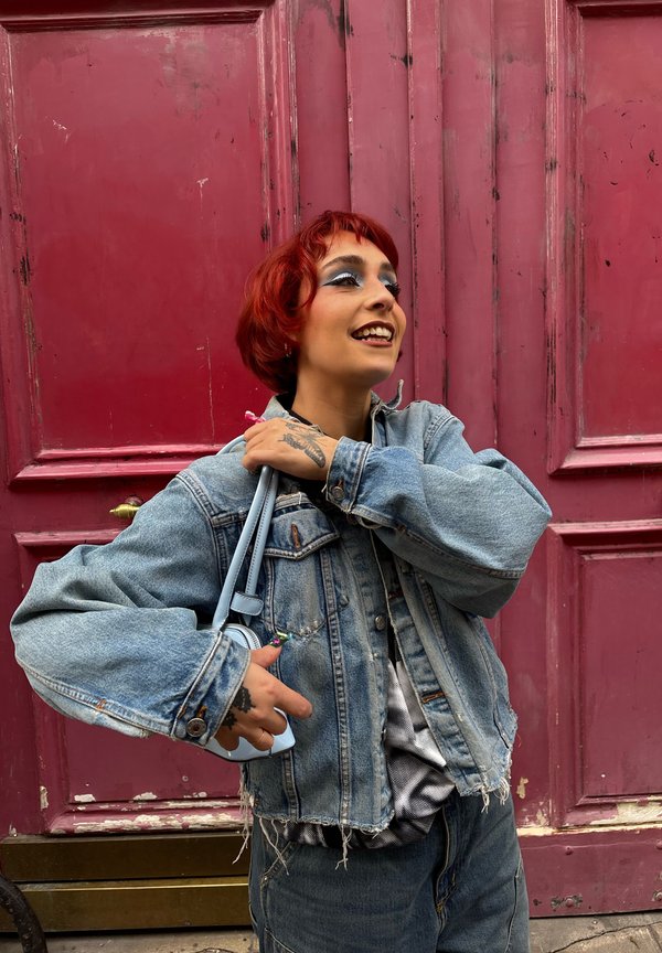 Ung person med kort rødt hår og makeup iført en slidt denimjakke, der holder en lille lyseblå pung, stående ved en rød dør.