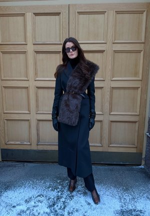 Mujer con un abrigo largo negro con una amplia estola de piel, guantes negros, gafas de sol y botas marrones, de pie sobre suelo nevado junto a unas puertas de madera.