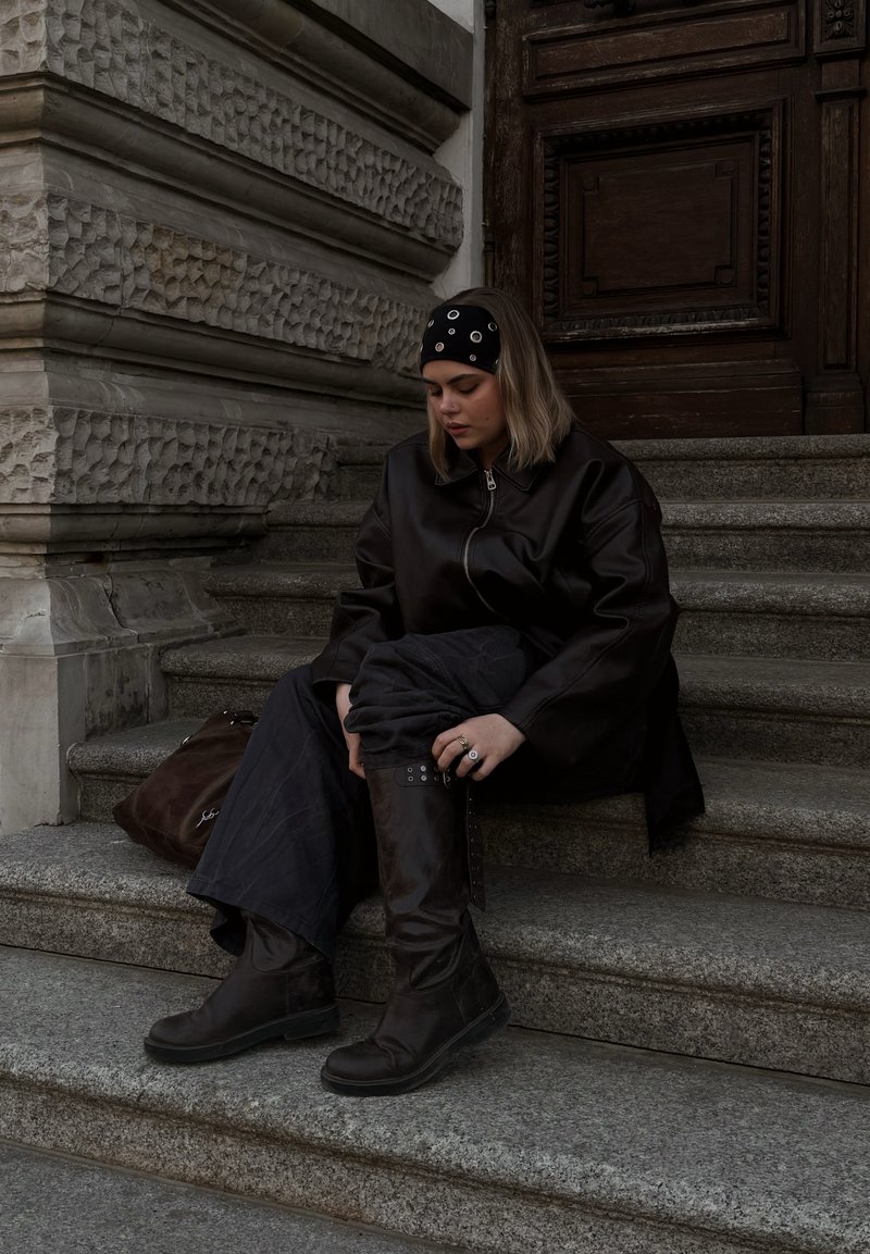 Jeune femme portant une veste en cuir noire et des bottes, assise sur des marches en pierre, ajustant sa botte, avec un grand sac marron à côté d'elle.