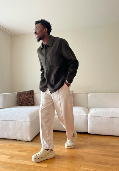 Hombre con pendientes sonriente, vestido con chaqueta oscura, pantalones claros y zapatos beige tipo slip-on, de pie en una sala de estar con sofá blanco y suelo de madera.