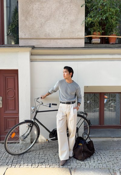 Bicicleta negra con herrajes plateados, suéter blanco y gris claro a rayas, pantalones anchos color crema y un bolso marrón oscuro en una calle de adoquines.