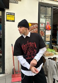 T-shirt noir oversize avec texte graphique rouge, superposé à une chemise blanche à col. Bonnet noir et bagues argentées. Décor urbain avec des étals de produits frais.