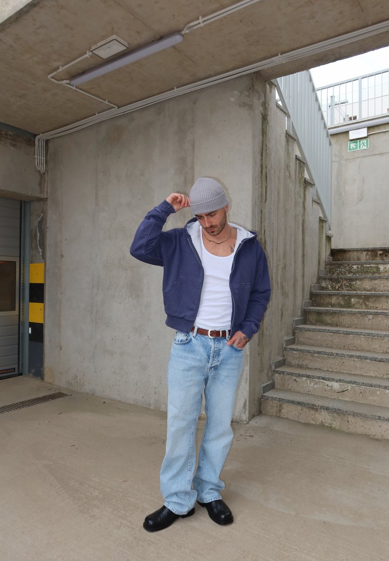 Man in een grijze muts, marineblauwe jas, wit hemd, lichte jeans en zwarte schoenen, staand in een betonnen trappenhuis naast de trap.