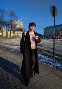 Personne portant un long manteau noir et une chemise rayée, tenant une tasse de café, marchant sur un trottoir en ville avec des bâtiments et des plaques de neige à proximité.