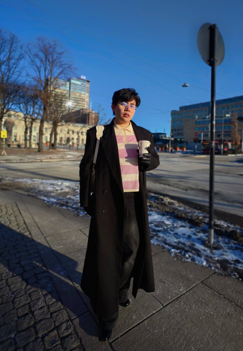 Personne portant un long manteau noir et une chemise rayée, tenant une tasse de café, marchant sur un trottoir en ville avec des bâtiments et des plaques de neige à proximité.