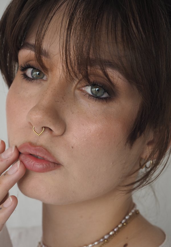 Junge Person mit kurzen braunen Haaren, grünen Augen, hellen Sommersprossen, goldenem herzförmigem Septumring und Perlenkette, die sanft ihre Lippen berührt.