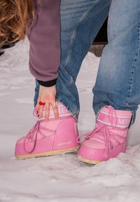 Personne portant des bottes de neige Moon Boot roses et un jean bleu, ajustant la botte sur un sol enneigé.