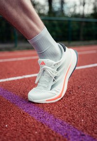 Valkoiset urheilukengät, joissa on verkkopinta, vaaleanpunaiset yksityiskohdat ja musta kanta; jalassa kevyet harmaat tekstuurimaiset sukat punaisella radalla, jossa on violetti viiva.