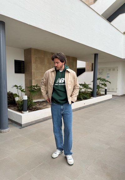 Chaqueta beige sobre sudadera verde, jeans azules y zapatillas blancas. De pie sobre un suelo de baldosas cerca de plantas, con una arquitectura moderna de fondo.