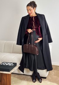Haut en velours bordeaux avec liens, jupe noire, long manteau noir, sac en cuir marron, et talons bordeaux ornés sur un canapé texturé.