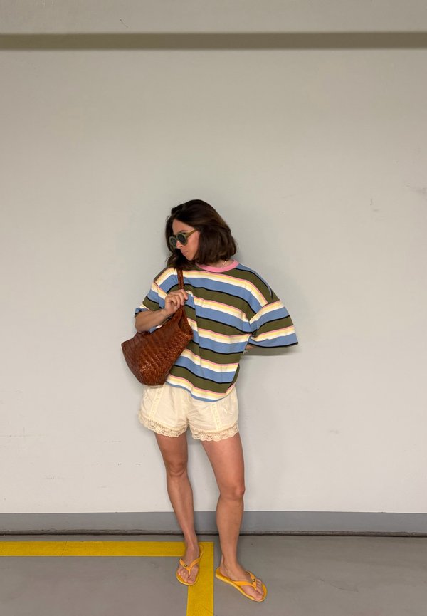 Femme portant des lunettes de soleil, un t-shirt à manches courtes rayé, un short crème, des tongs jaunes, tenant un sac à main tressé marron contre un mur uni.