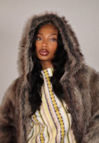 Manteau à capuche en fausse fourrure brun foncé avec une finition douce et texturée. Porté sur un haut rayé aux lignes jaunes, noires et blanches.