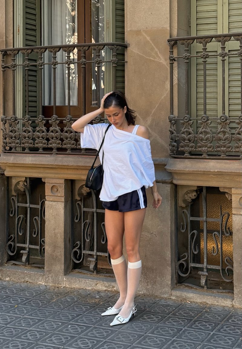 Femme en chemise blanche épaules dénudées, short noir, chaussettes blanches montantes et talons, debout sur un pavement à motifs près d'une balustrade de balcon ornée.