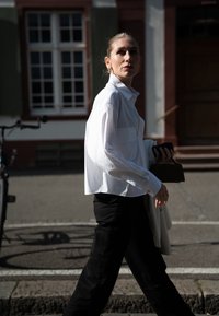 Balta marškiniai su trumpu dizainu ir dviem priešais esantys kišenėmis. Suderinta su juodomis kelnėmis, laikoma maža rankinė ir telefonas. Saulės apšviesta aplinka.
