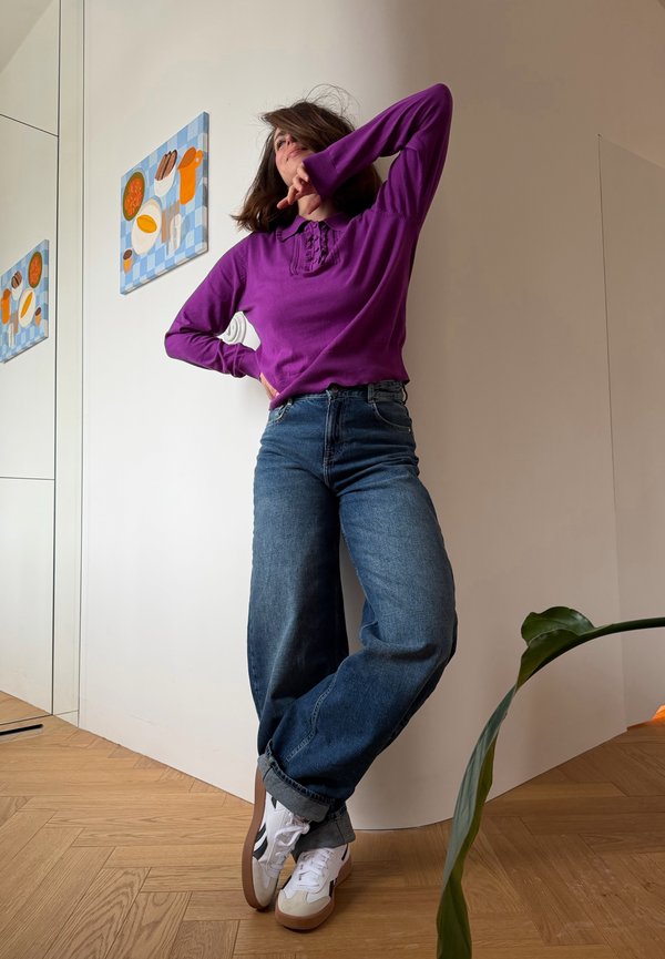 Osoba v fialovom tričku a širokých džínsach pózuje s jednou rukou ohnutou pri tvári v moderne zariadenej miestnosti s drevenou podlahou a nástenným umením.