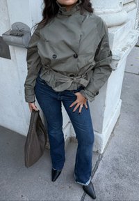 Jachetă verde măslinie rezistentă la apă, cu design cu nasturi dubli, talie cu curea și guler înalt. Asortată cu blugi evazați din denim închis la culoare și pantofi cu vârf ascuțit.