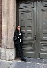 Blazer nero, camicia bianca, pantaloni neri e sneakers. Appoggiato contro un muro di pietra testurizzato accanto a una grande porta verde a pannelli.