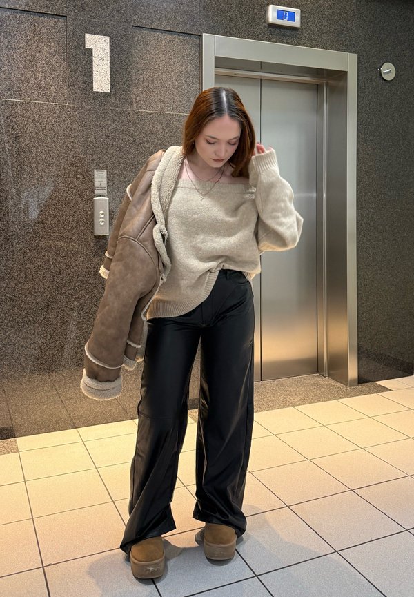 Donna in piedi vicino all'ascensore, indossa un maglione beige, pantaloni neri, scarpe marroni e tiene una giacca marrone sul braccio sinistro.
