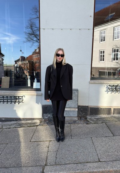Blazer oversize negro sobre un cuello de tortuga negro, combinado con leggins negros brillantes y botas de tacón alto hasta la rodilla. Las gafas de sol completan el look.