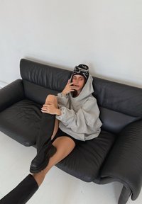 Šedá oversized mikina s kapucí, vysoké černé boty, vzorovaná černá čepice. Sedí na černé kožené pohovce, za ní je jednoduchá bílá zeď.