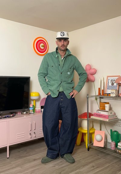 Modelo con una chaqueta verde de pana sobre una camisa más clara y vaqueros azules de gran tamaño. La habitación incluye decoración y muebles coloridos.
