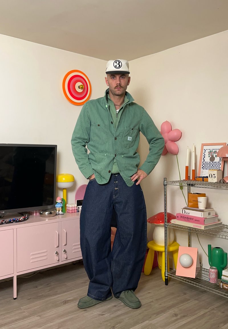 Modello che indossa una giacca verde in velluto a coste sopra una camicia più chiara e jeans blu oversize. La stanza è arredata con decorazioni e mobili colorati.