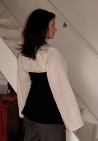 Top preto ajustado colocado por baixo de uma jaqueta oversized creme com riscas e mangas largas, combinado com calças cinzentas; decoração em madeira e metal ao fundo.