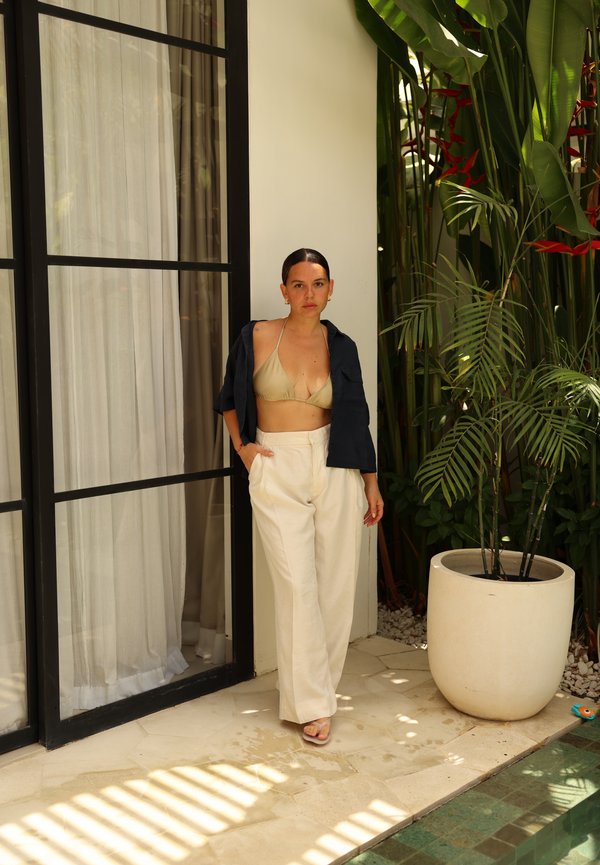Mujer de pie junto a una puerta de cristal, vistiendo un top de bikini beige, pantalones blancos de cintura alta y una camisa negra abierta, junto a una gran planta tropical en maceta.