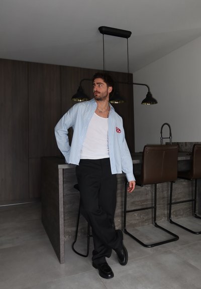 Hombre con camisa de rayas y pantalones negros apoyado en la isla de la cocina con taburetes de bar y lámparas colgantes modernas.